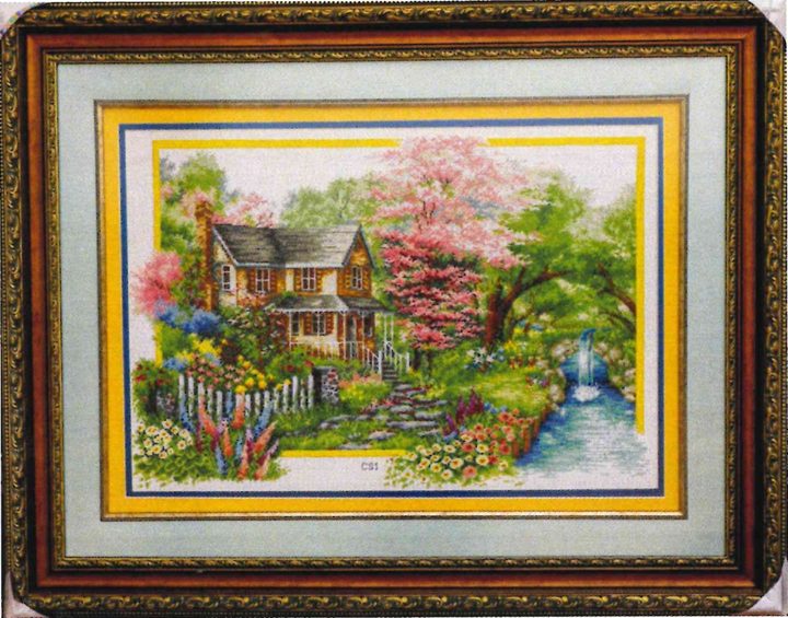 Cross-stitch artwork of a cottage among blossoming trees, with a picket fence, flower beds and a small waterfall, in a carved frame.