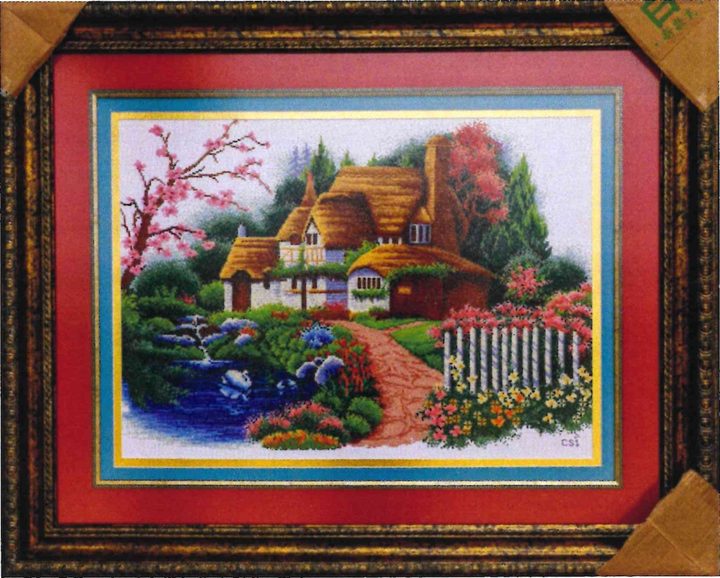 Cross-stitch artwork of a thatched cottage beside a pond with swans, pink blossom and a flowering garden path, on a red mat in an ornate frame.