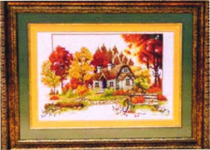 Cross-stitch artwork of a cottage surrounded by autumn trees in red, orange and gold, in a wooden frame with a green mat.