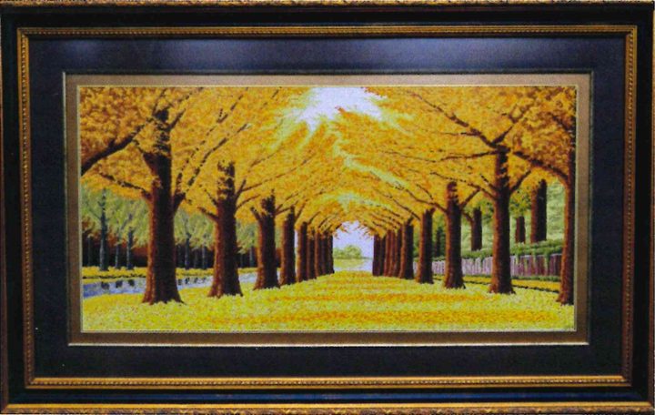 Cross-stitch artwork of a golden tree-lined avenue beside water, stitched in yellows and browns, in a black and gold frame.