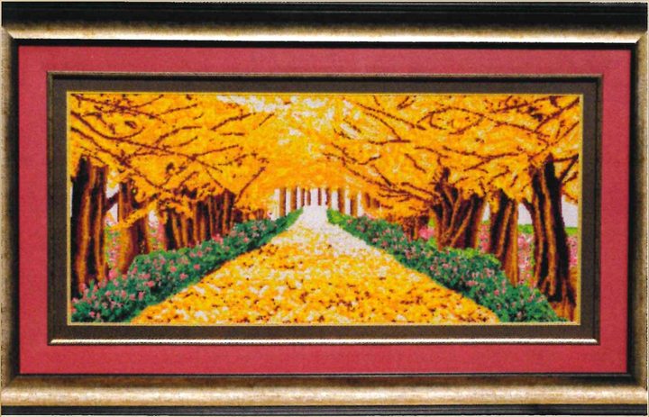 Cross-stitch artwork of a path carpeted in yellow leaves between autumn trees and pink flowers, on a red mat in a gold frame.