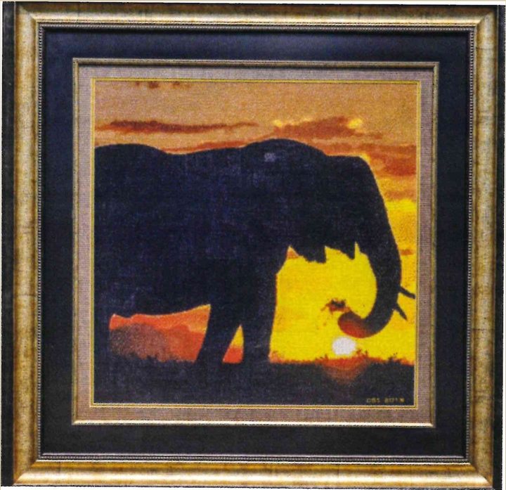 Cross-stitch artwork of an elephant in black silhouette against an orange and yellow sunset, trunk curled towards the setting sun, in a gold frame.
