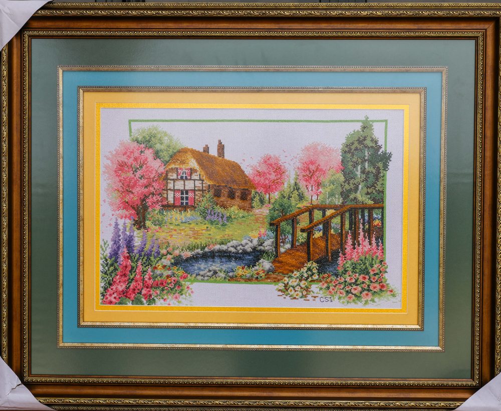Framed cross stitch of a cottage with a garden bridge and blossom trees.