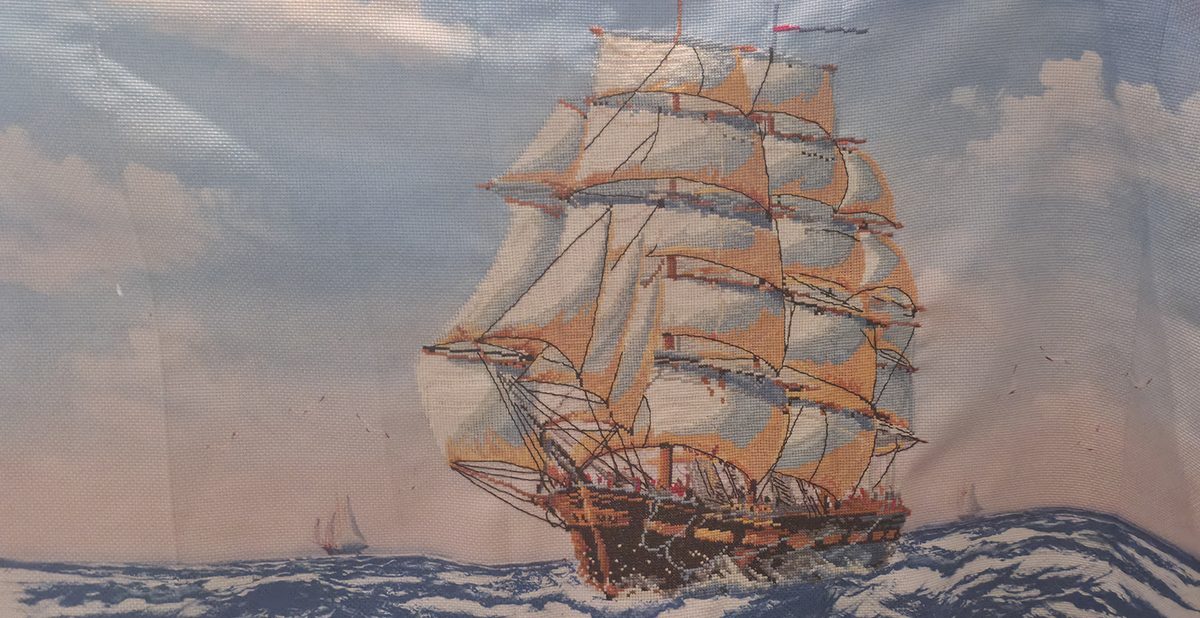 Cross stitch of a tall ship under full sail on a rolling blue sea, shown before framing.