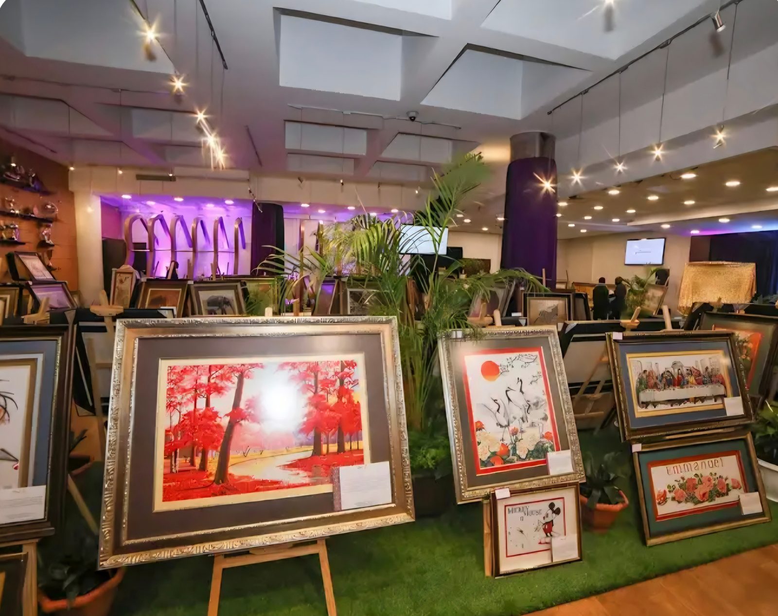 Framed cross-stitch artworks on easels at an indoor exhibition, including a red forest scene, cranes and The Last Supper.