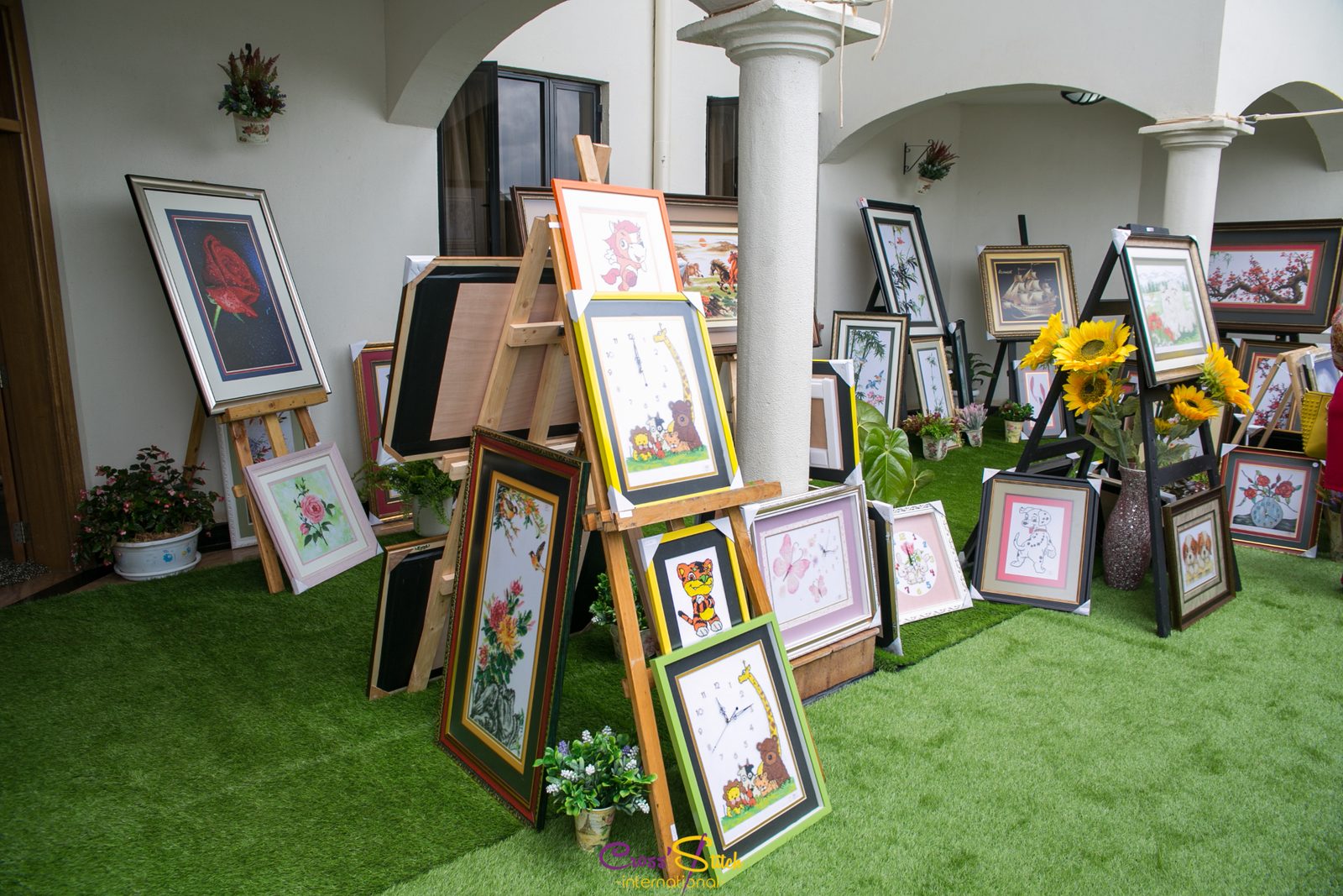 Framed cross-stitch artworks displayed on easels and against the walls of a veranda, with potted sunflowers and plants.