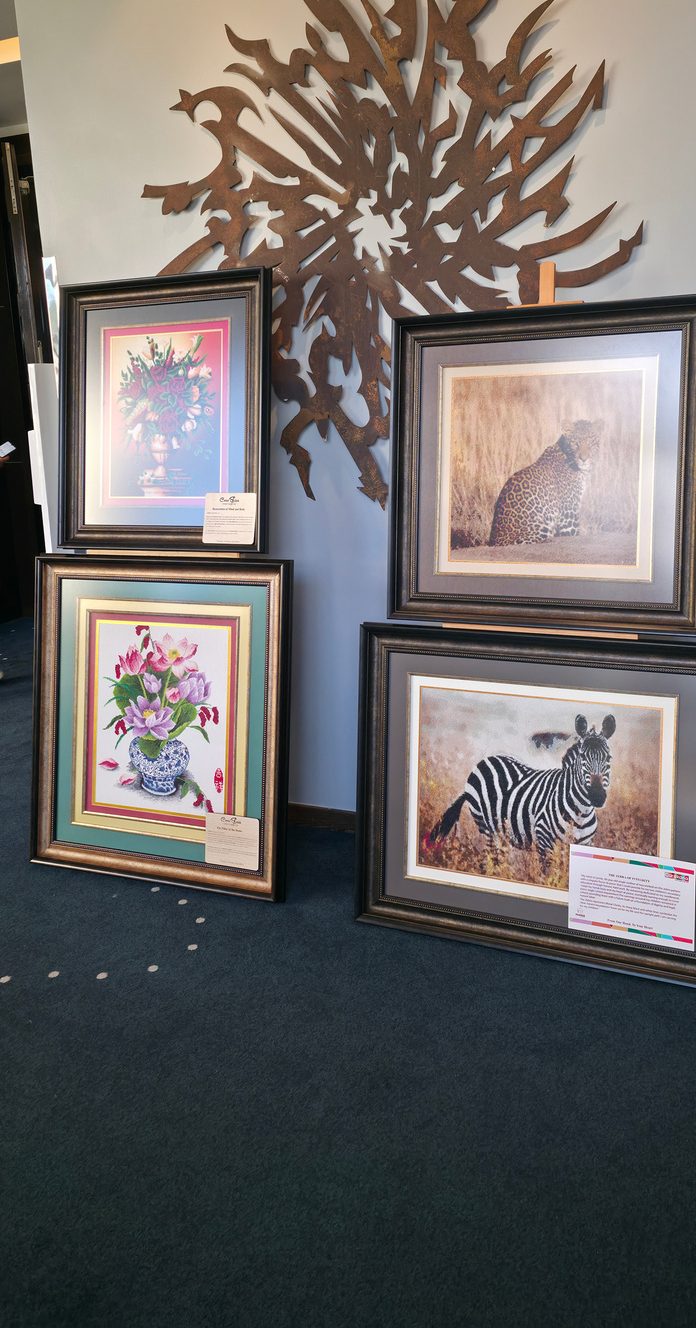 Four framed cross-stitch pieces, a leopard, a zebra and two flower studies, standing against a wall on display.