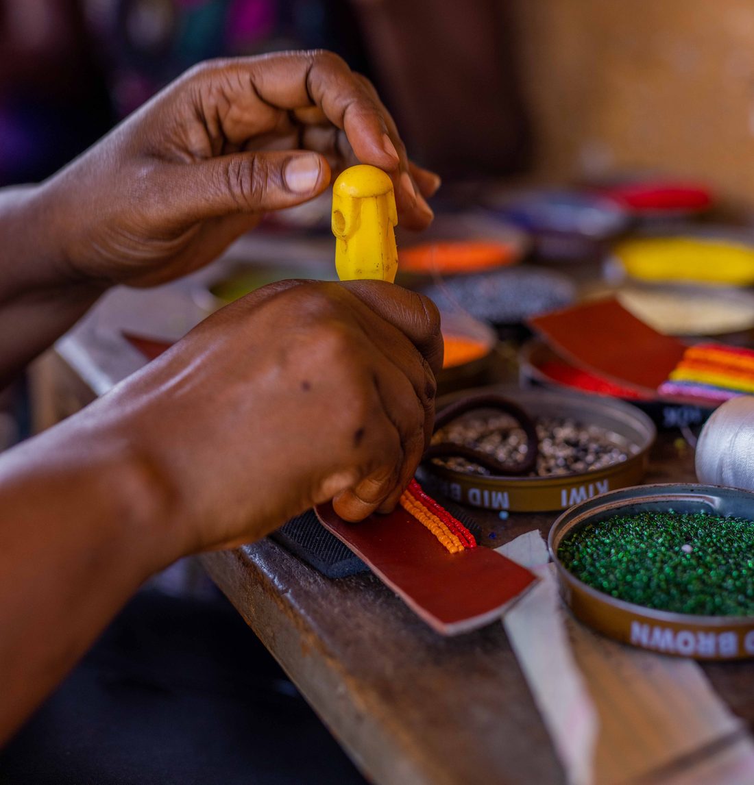 An artisan's hands threading coloured beads at a workbench scattered with tins of beads.