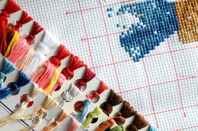 Skeins of coloured embroidery thread on a numbered organiser beside evenweave fabric marked with a red grid, part-stitched in blue and gold.